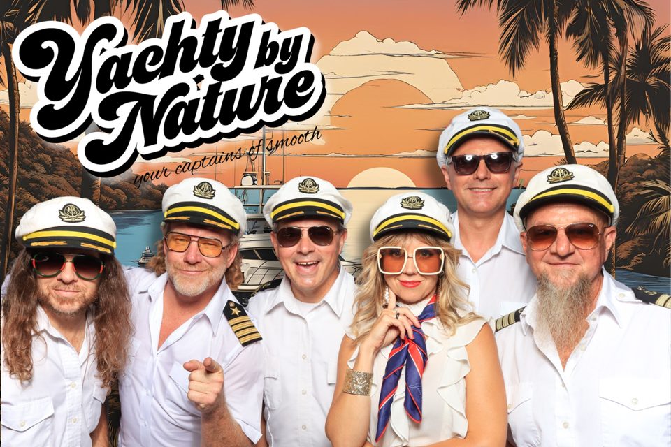 Yachty By Nature in captain uniforms, photographed against palms, a yacht, and a sunset.