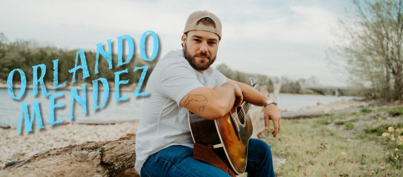 Orlando Mendez seated outdoors with an acoustic guitar, his name in blue lettering beside him.